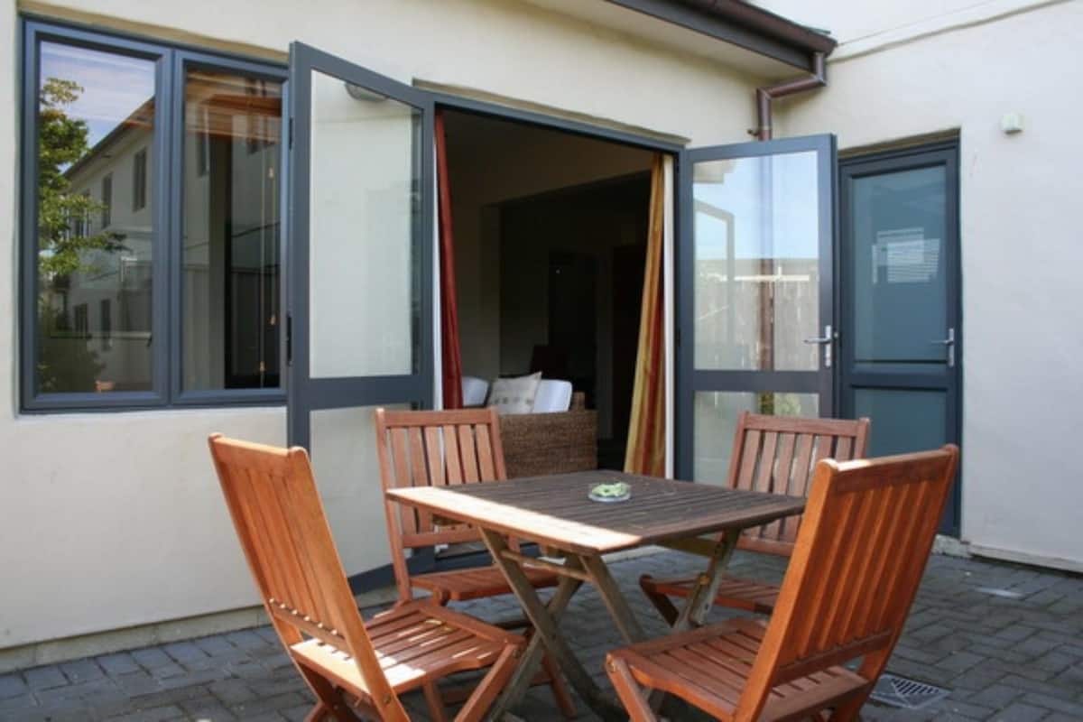 An open patio with a wooden table and chairs, a wicker sofa, and a sliding glass door leading to a room.
