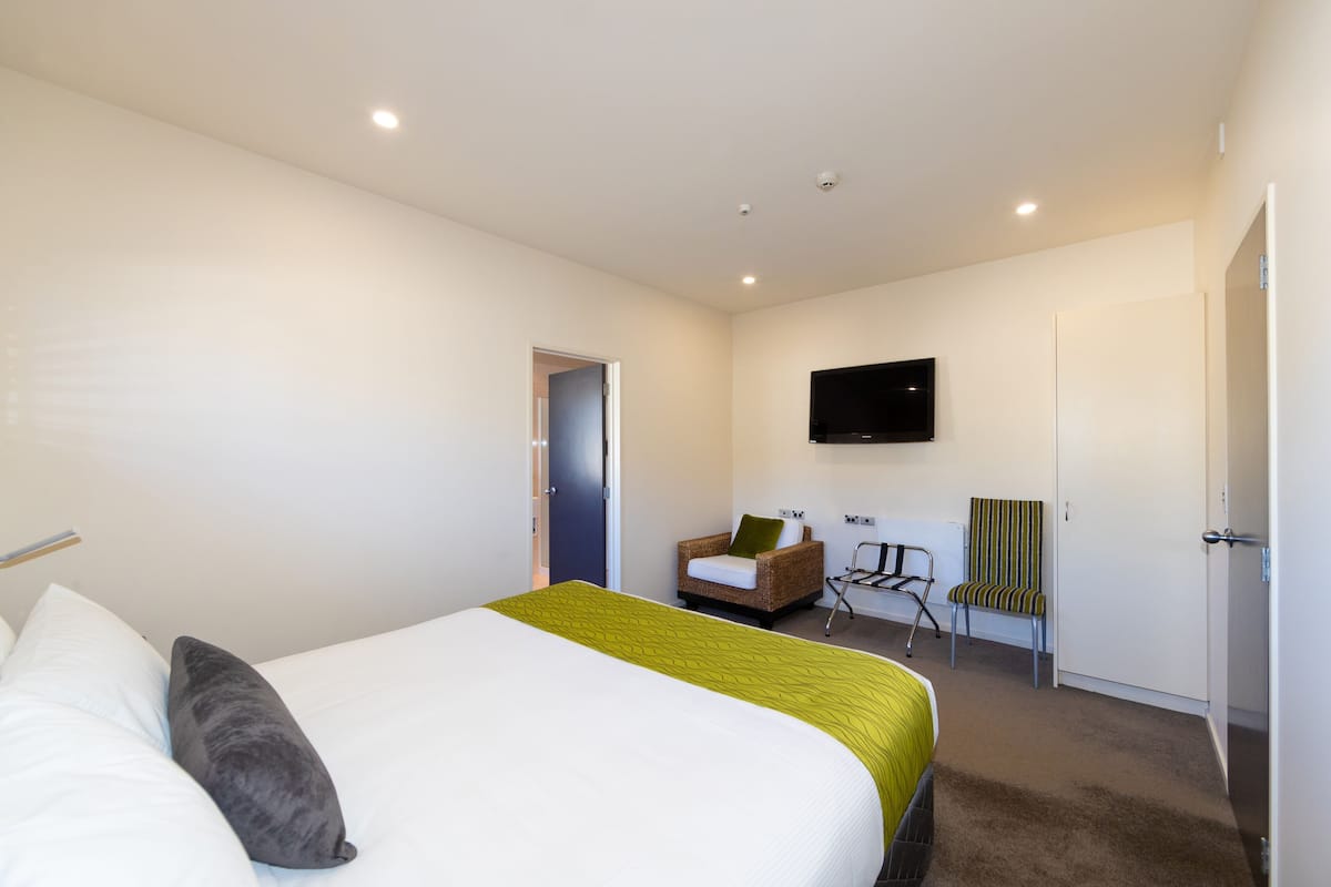 A modern hotel room with a large bed, a flat-screen TV mounted on the wall, a wicker chair, and a desk with a chair.