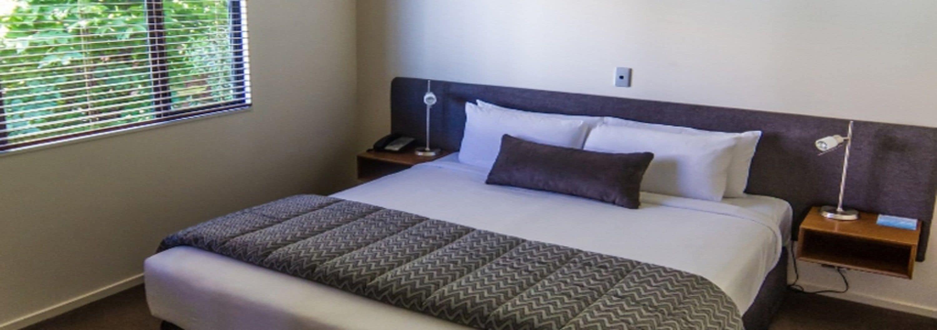 Una cama bien tendida con una colcha estampada, una lámpara de noche y una mesita de noche con un teléfono.