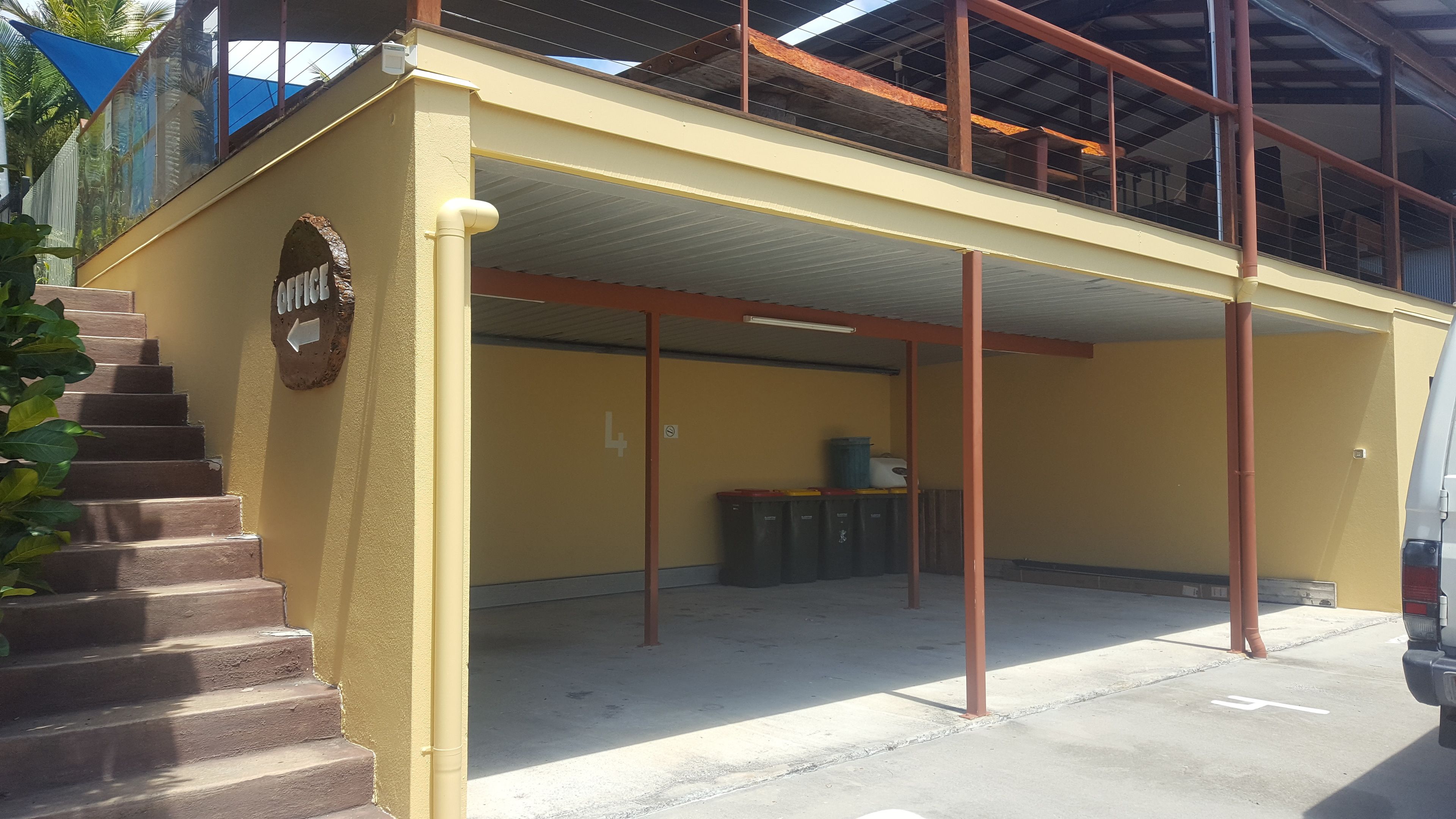 Un edificio giallo con una scala a sinistra, una zona coperta con un cestino e un veicolo parcheggiato davanti.