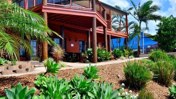A modern two-story building with a balcony, surrounded by tropical plants and a clear blue sky.