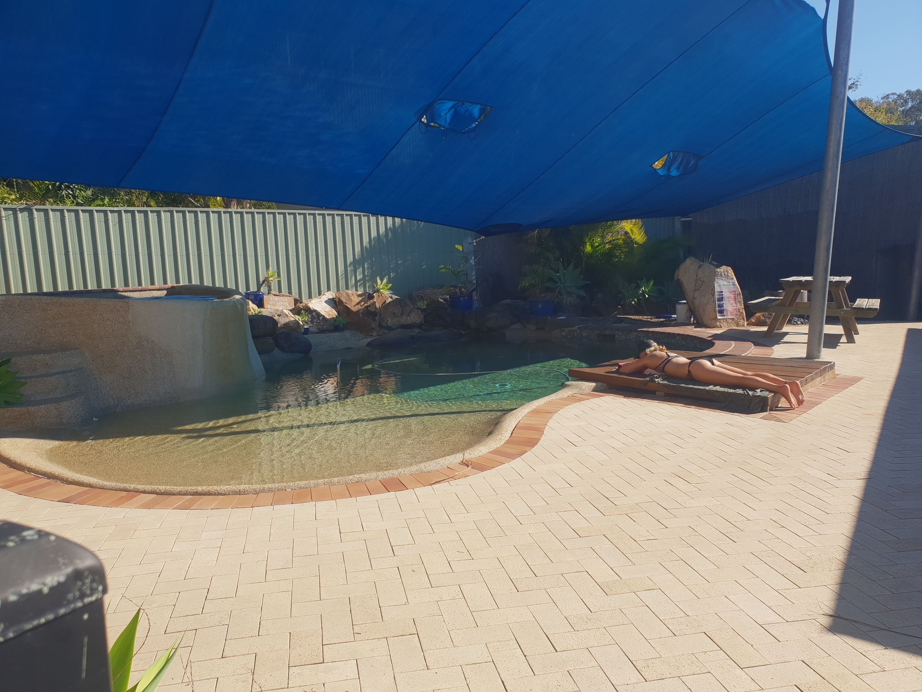 A pool area with a blue canopy, a brick patio, and a wooden deck.