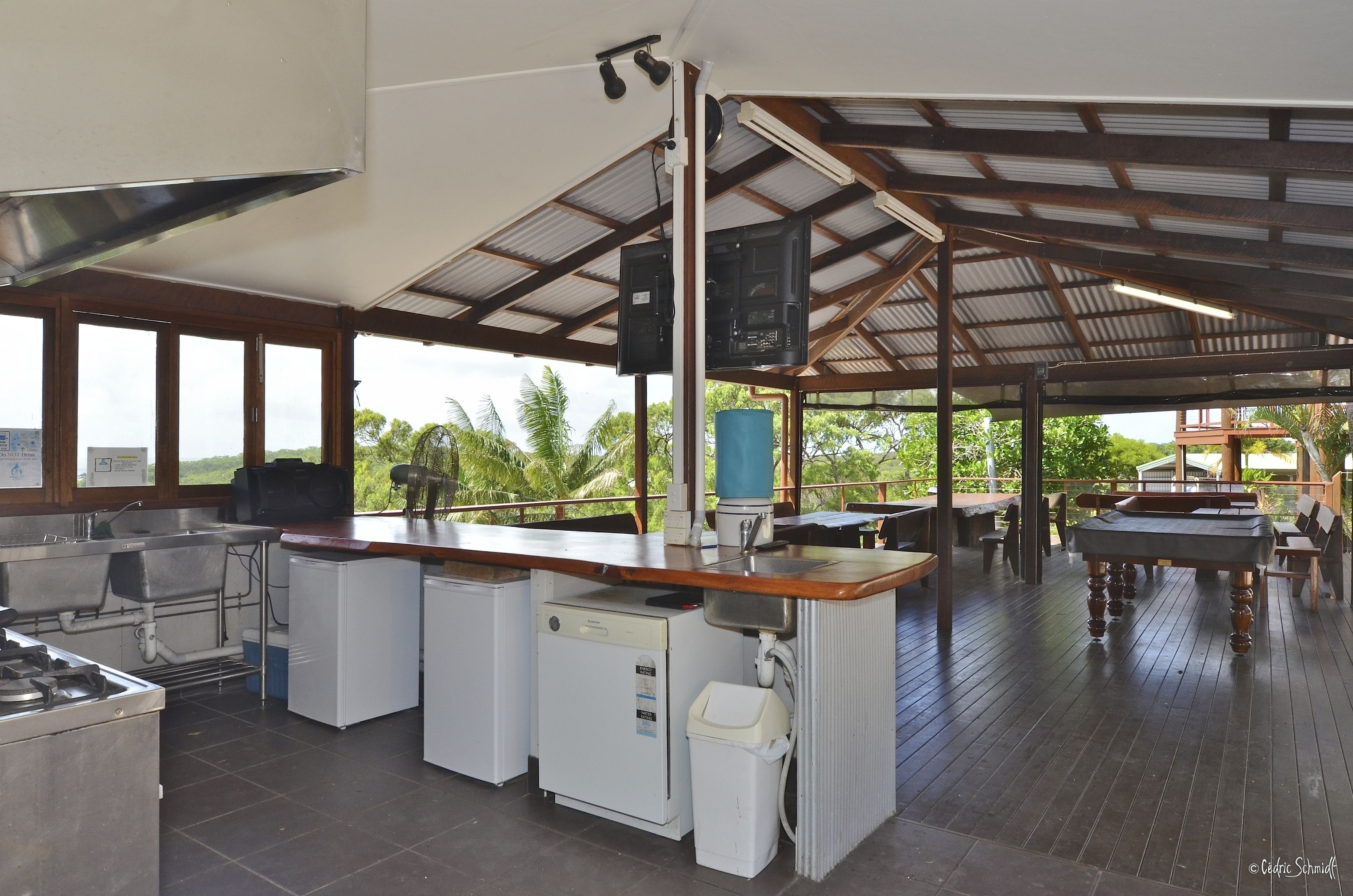 Un área de cocina exterior amplia, con una parrilla, pileta y refrigerador, rodeada de mesas y sillas de madera.