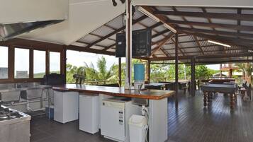Un área de cocina exterior amplia, con una parrilla, pileta y refrigerador, rodeada de mesas y sillas de madera.