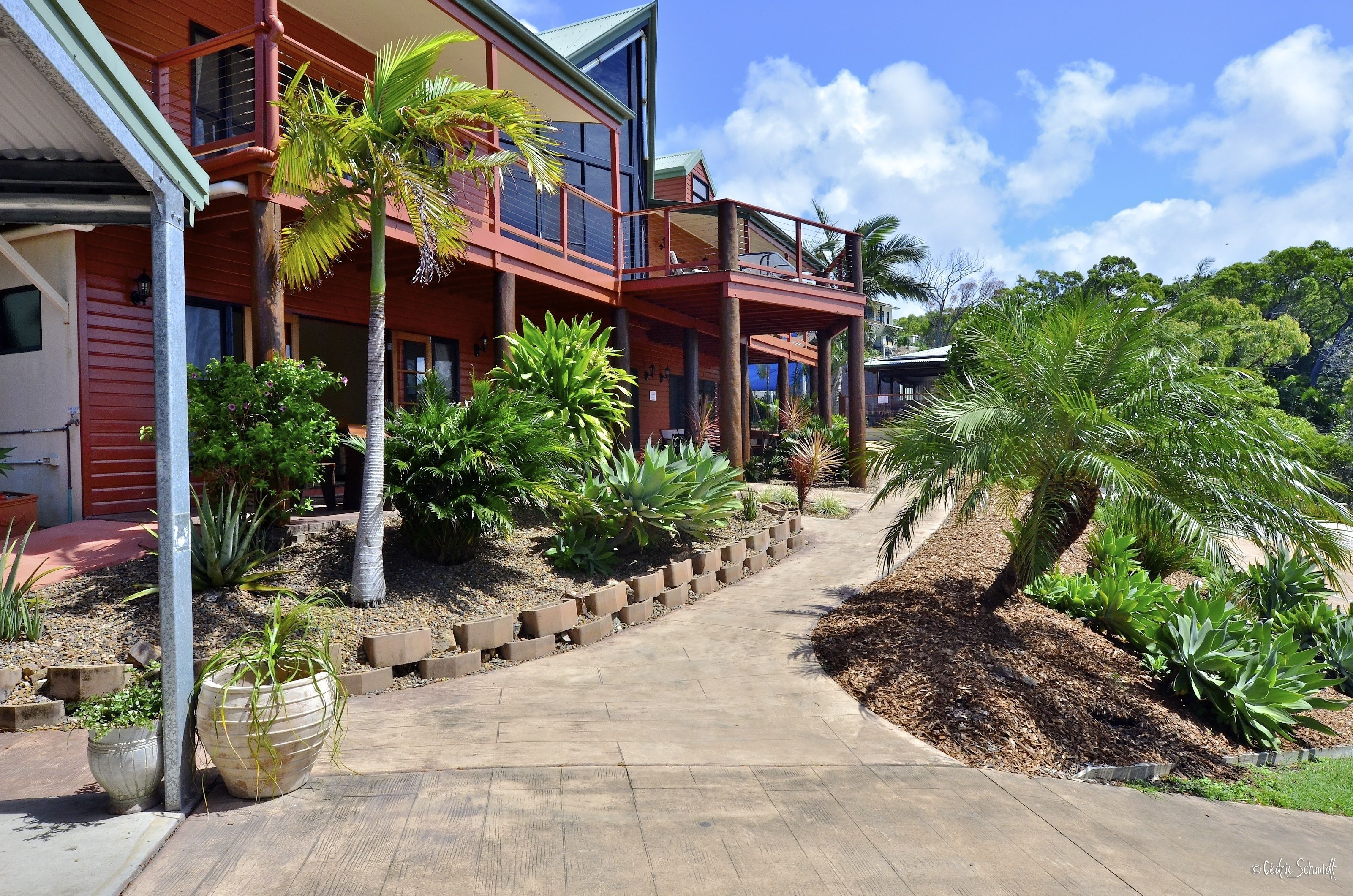 A modern two-story building with a balcony, surrounded by tropical plants and a paved walkway.
