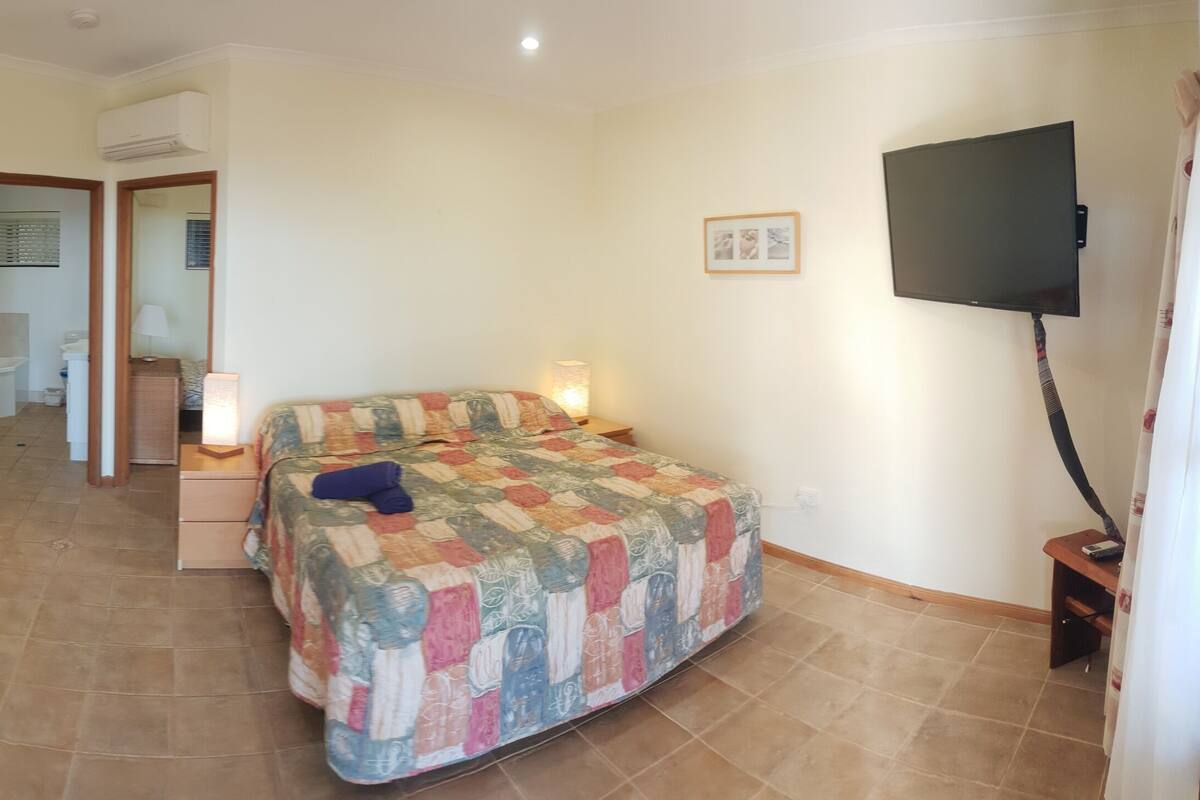 A compact hotel room with a sofa, a small wooden table, a bed with a colorful quilt, a desk with a computer, and a TV mounted on the wall.