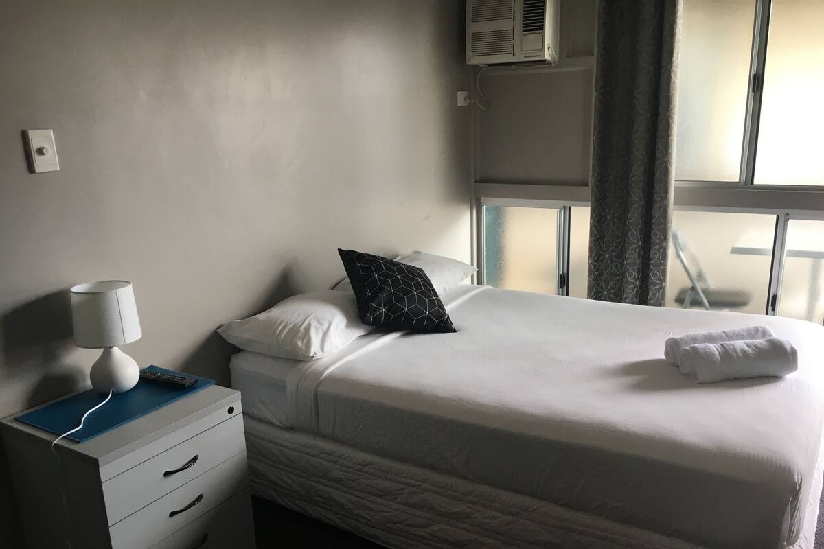 A hotel room with a bed, a bedside table with a lamp, a dresser, and a window with curtains.