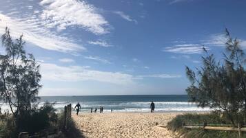 A sandy beach with a wooden fence and trees on the left, and a clear blue sky with scattered clouds.