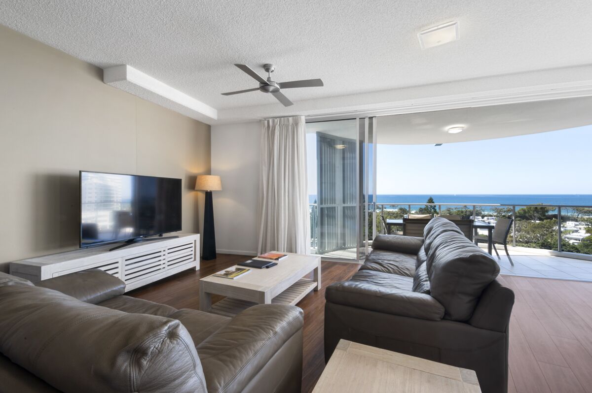 A modern living room with a flat-screen TV, leather sofas, a coffee table, and a balcony with a view of the ocean.