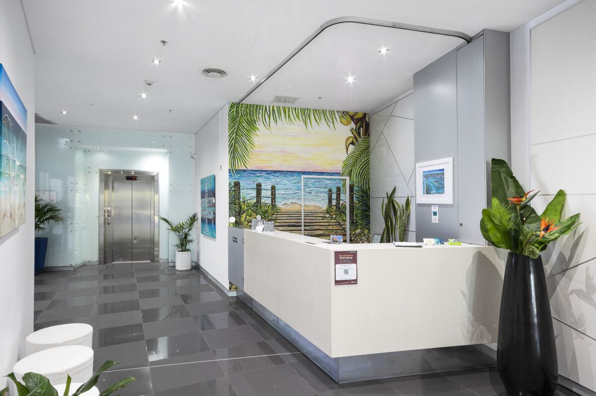 A reception area with a check-in counter, a reception desk, a wall mural of a beach, and a potted plant.