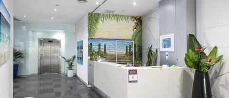 A reception area with a check-in counter, a reception desk, a wall mural of a beach, and a potted plant.