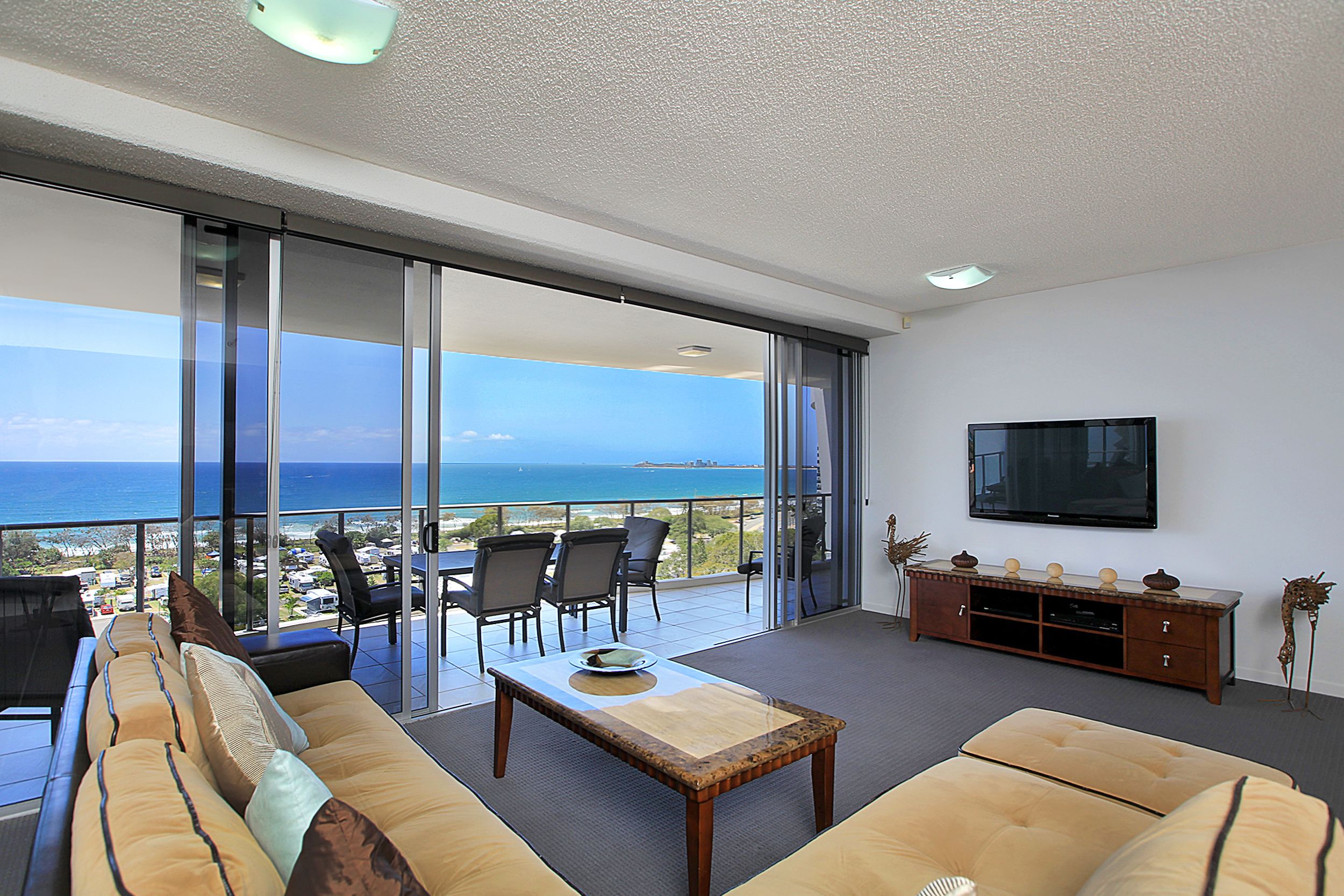 Ampla sala de estar com grande porta de vidro deslizante que dá para uma varanda com vista para o oceano. Há um sofá, uma mesa de centro e uma TV fixada na parede.