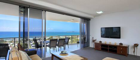 Ampla sala de estar com grande porta de vidro deslizante que dá para uma varanda com vista para o oceano. Há um sofá, uma mesa de centro e uma TV fixada na parede.