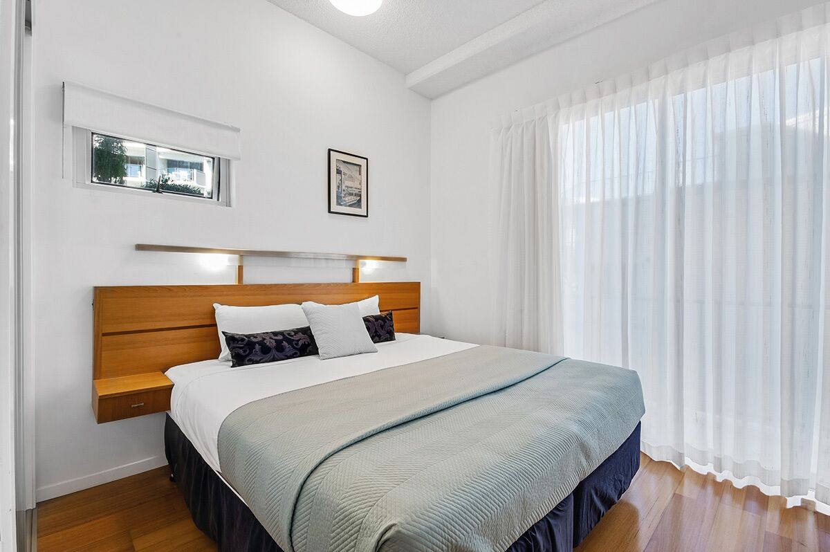 A neatly arranged bedroom with a wooden headboard, a bed with white and patterned pillows, a bedside table, and a window with a view of greenery.