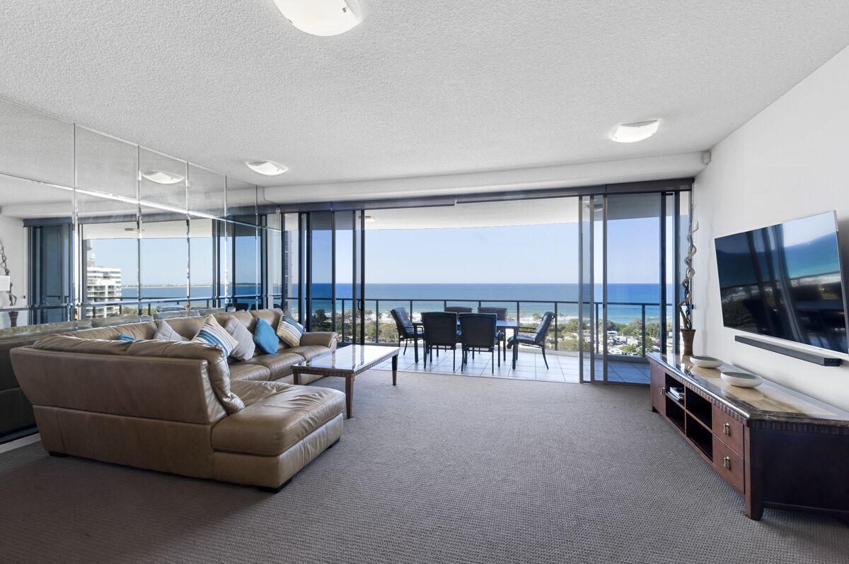 A modern living room with a large sofa, a coffee table, and a TV on a stand. There is a balcony with a view of the ocean.