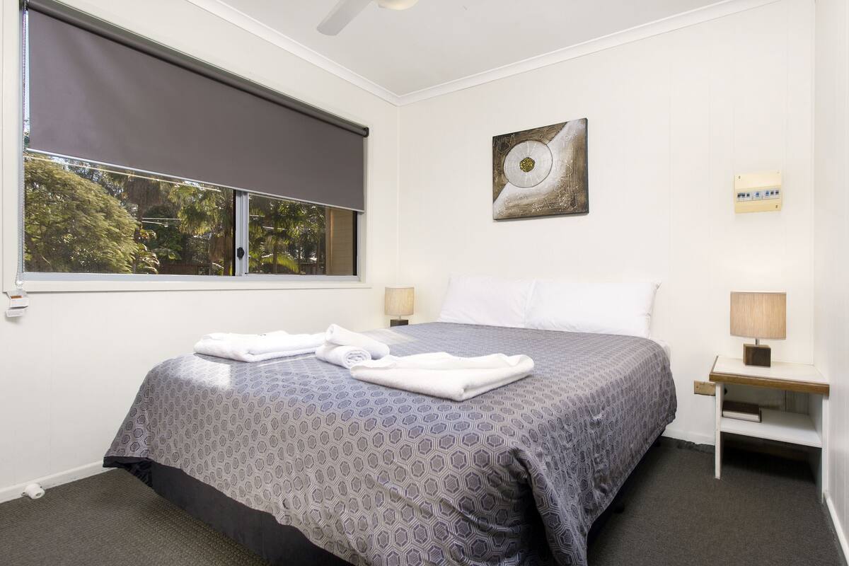 A neatly made bed with a patterned bedspread, a window with a blind, a wall-mounted artwork, and a bedside table with a lamp.