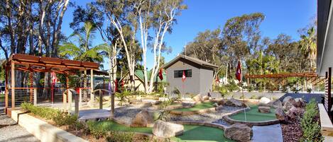 A miniature golf course with a small building, trees, and a wooden pergola.