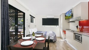 Habitación compacta con mesa de comedor para dos, cama con televisor encima, kitchenette y puerta corrediza de vidrio que da a un espacio exterior.