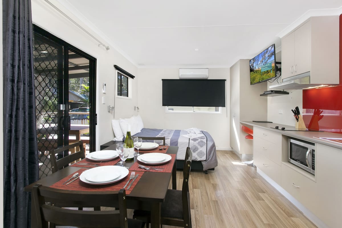 A compact room with a dining table set for two, a bed with a TV above it, a kitchenette, and a sliding glass door leading to an outdoor area.