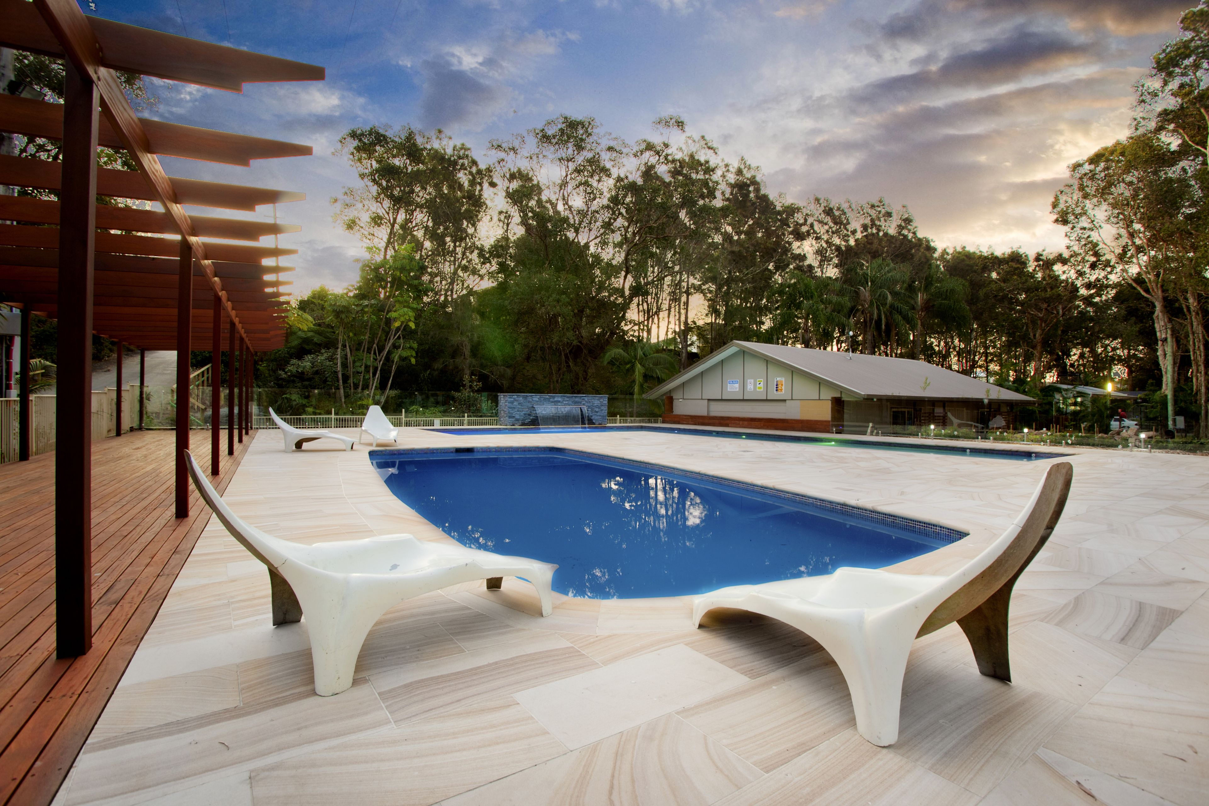 A swimming pool with two white lounge chairs, a wooden deck, and a view of trees and a building in the background.