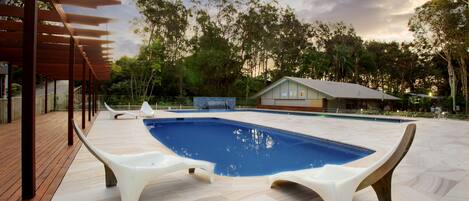A swimming pool with two white lounge chairs, a wooden deck, and a view of trees and a building in the background.