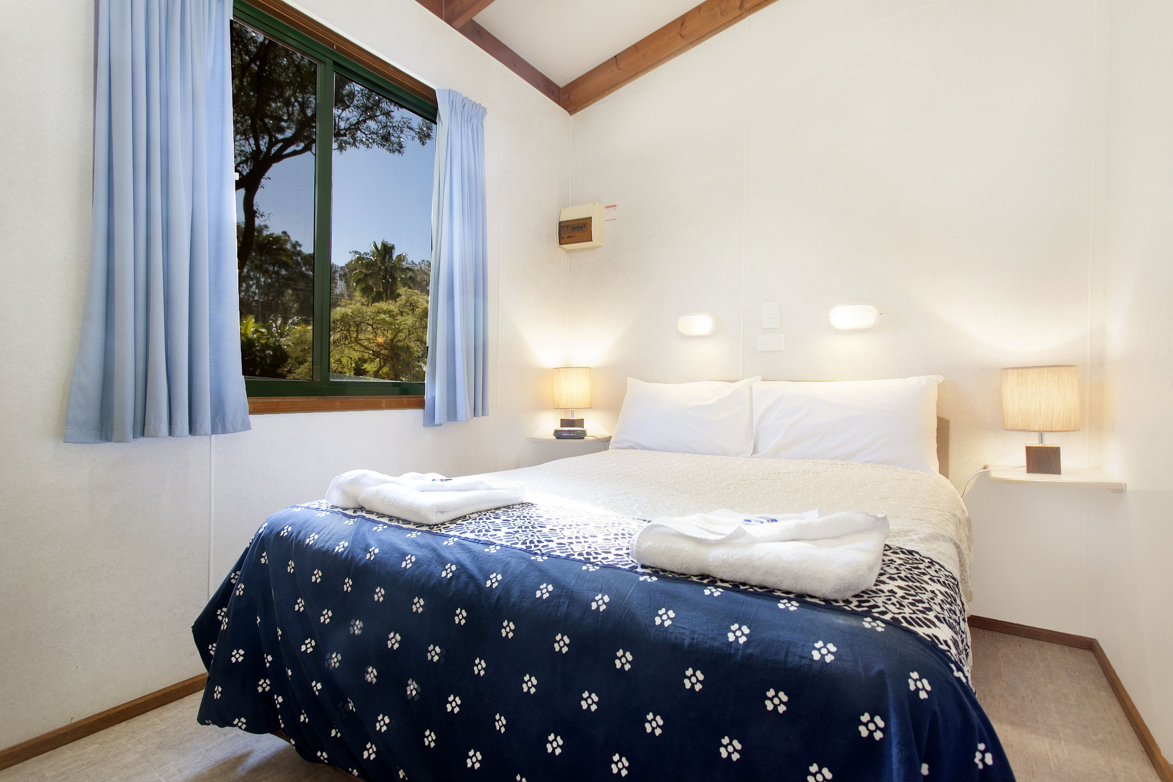 A neatly made bed with a blue floral bedspread, two bedside lamps, and a window with curtains.