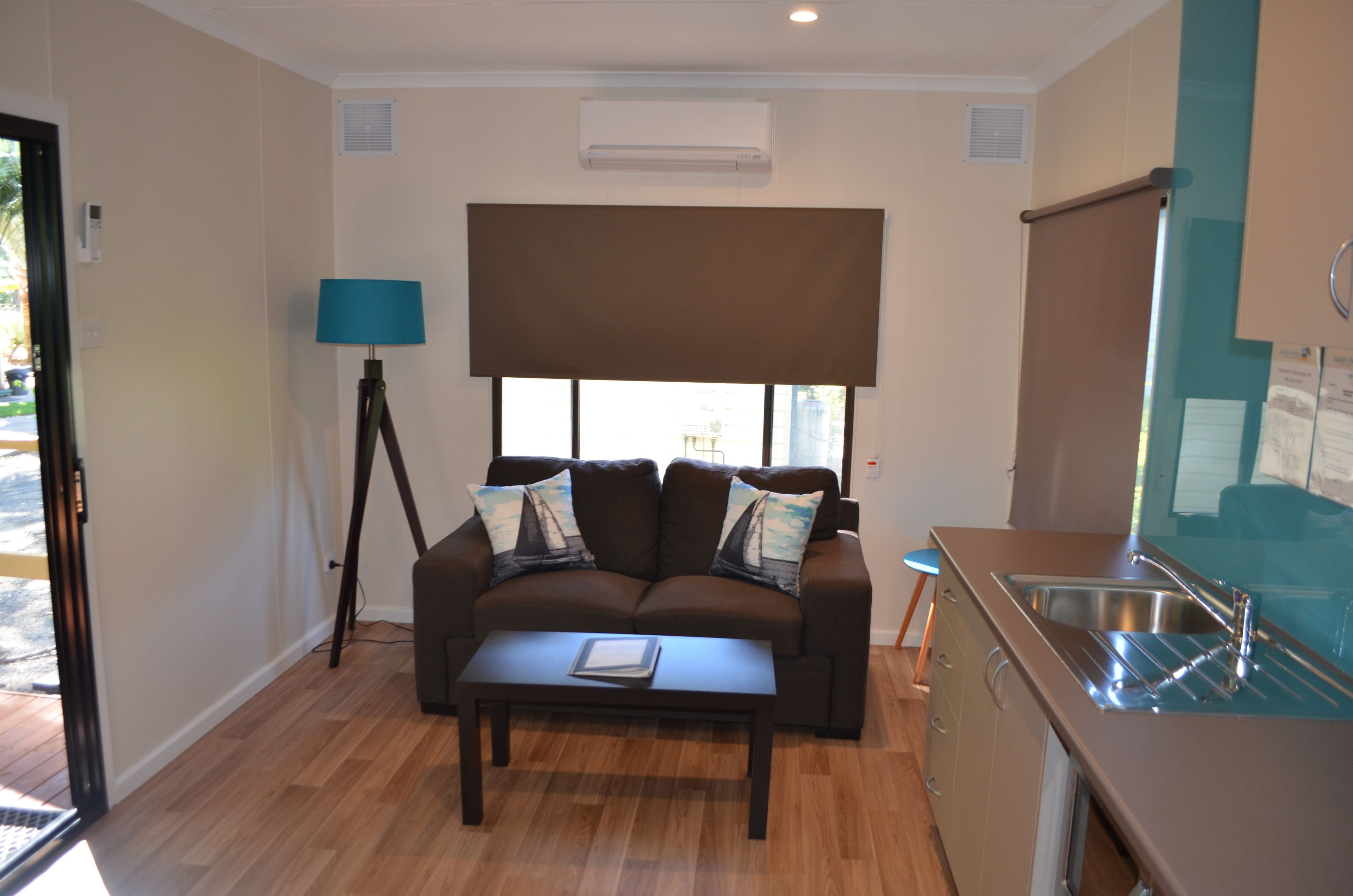 A compact living space with a sofa, a coffee table, a lamp, a window with a blind, and a kitchenette with a sink.
