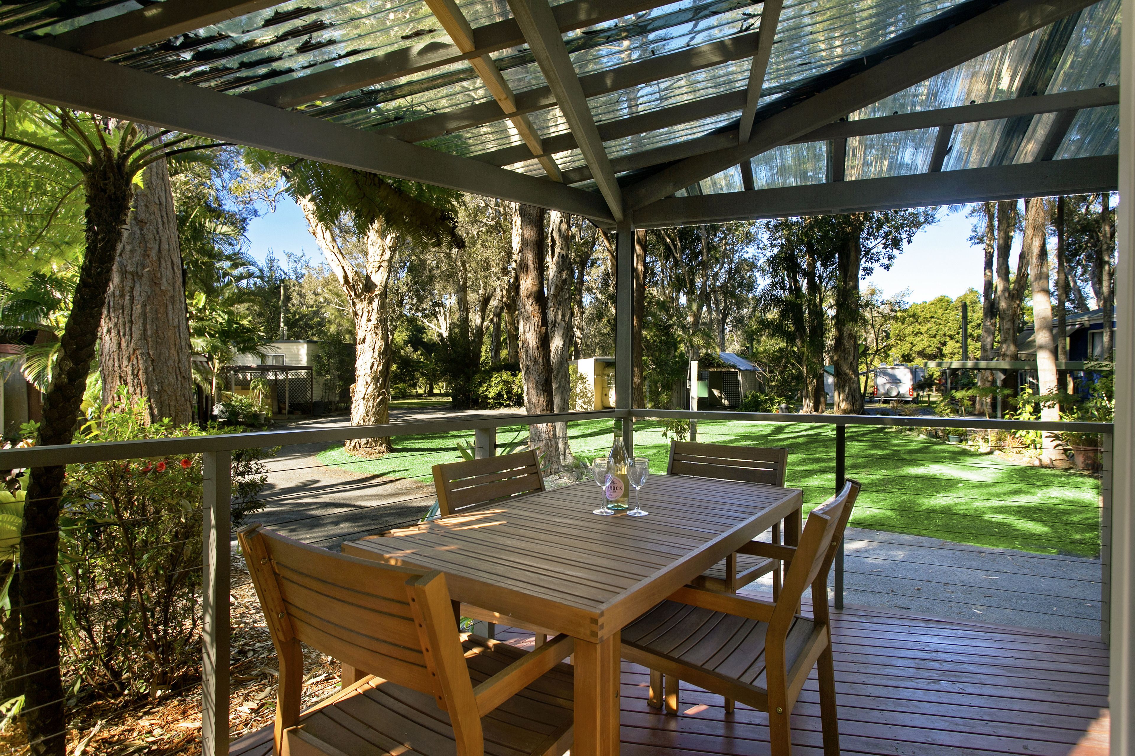 A wooden deck with a table and chairs, surrounded by trees and a grassy lawn.