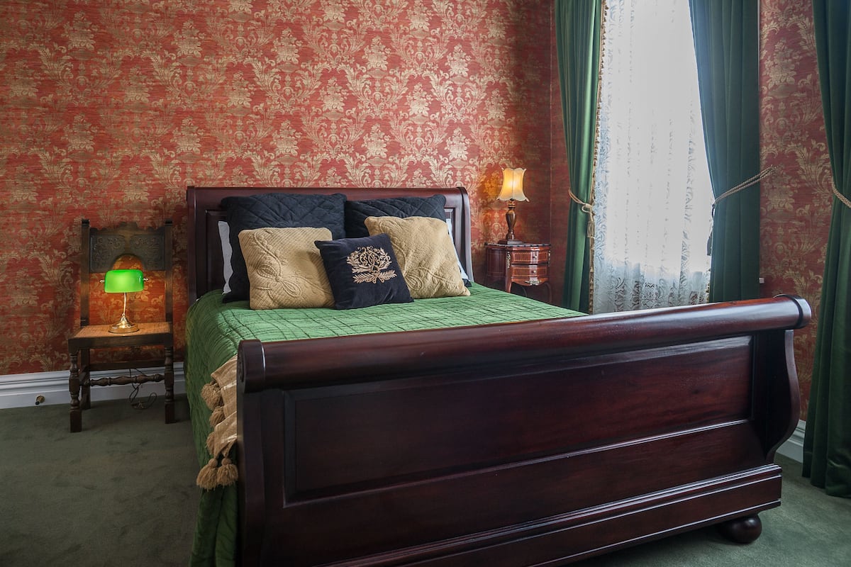 Una cama con dosel tradicional, con cubrecama verde y varios almohadones, una mesita de noche con lámpara verde y pared cubierta de papel tapiz con motivos.