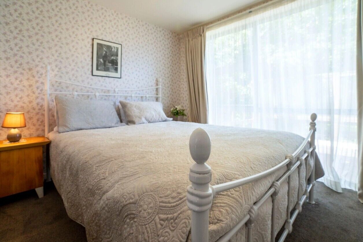A neatly made bed with a white headboard and footboard, a bedside table with a lamp, and a window with sheer curtains.