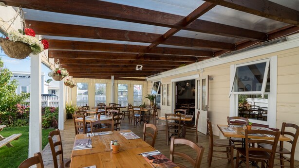 A covered patio area with wooden tables and chairs, a white fence, and a view of a house in the background.