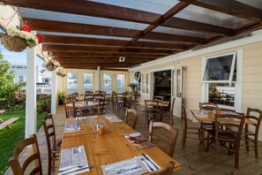 Ein überdachter Patio-Bereich mit Holztischen und -stühlen, einem weißen Zaun und Blick auf ein Haus im Hintergrund.