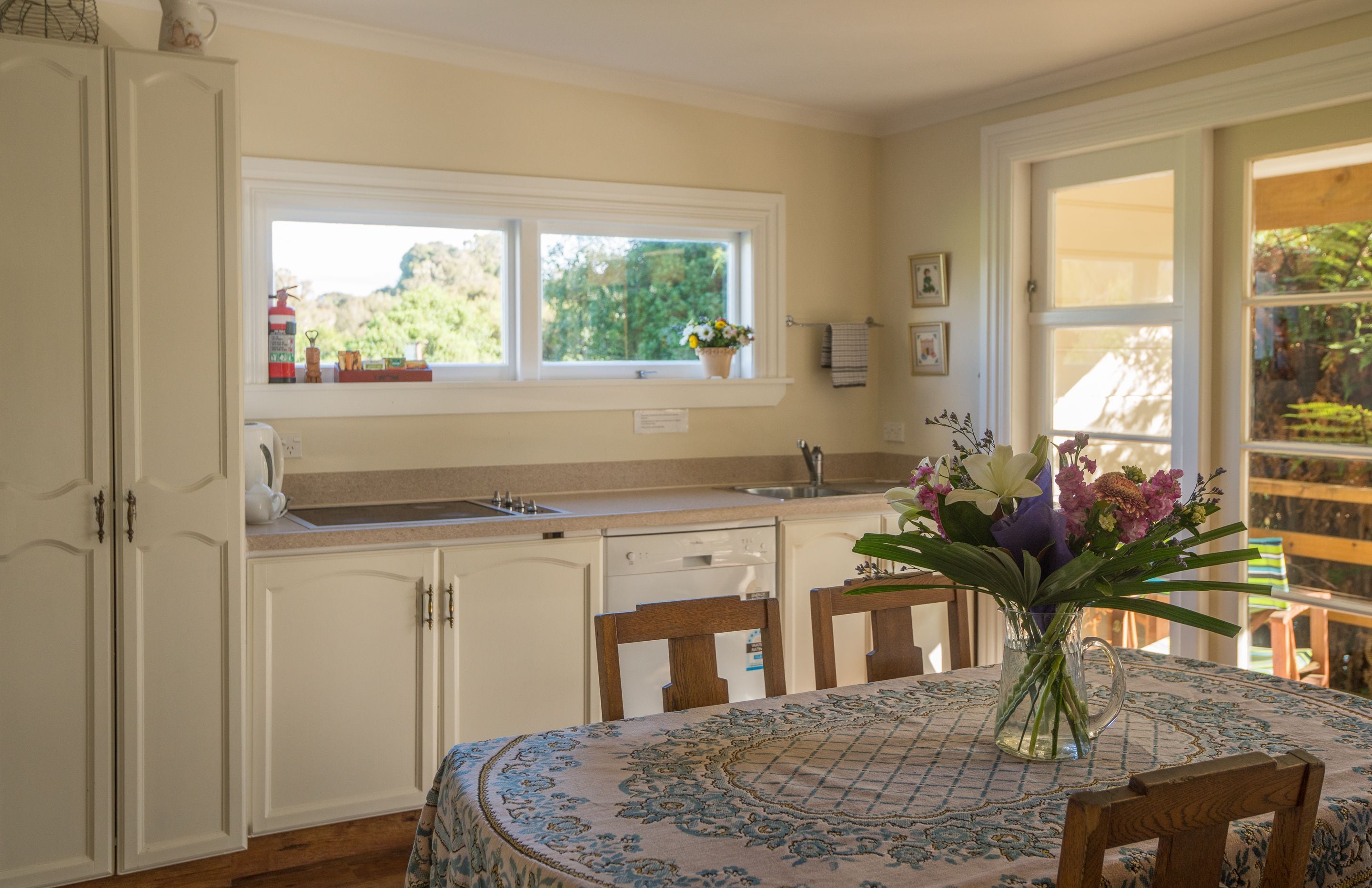 Una cocina con armarios blancos, una ventana con una maceta y una mesa de comedor con mantel floral.