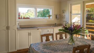 Una cocina con armarios blancos, una ventana con una maceta y una mesa de comedor con mantel floral.