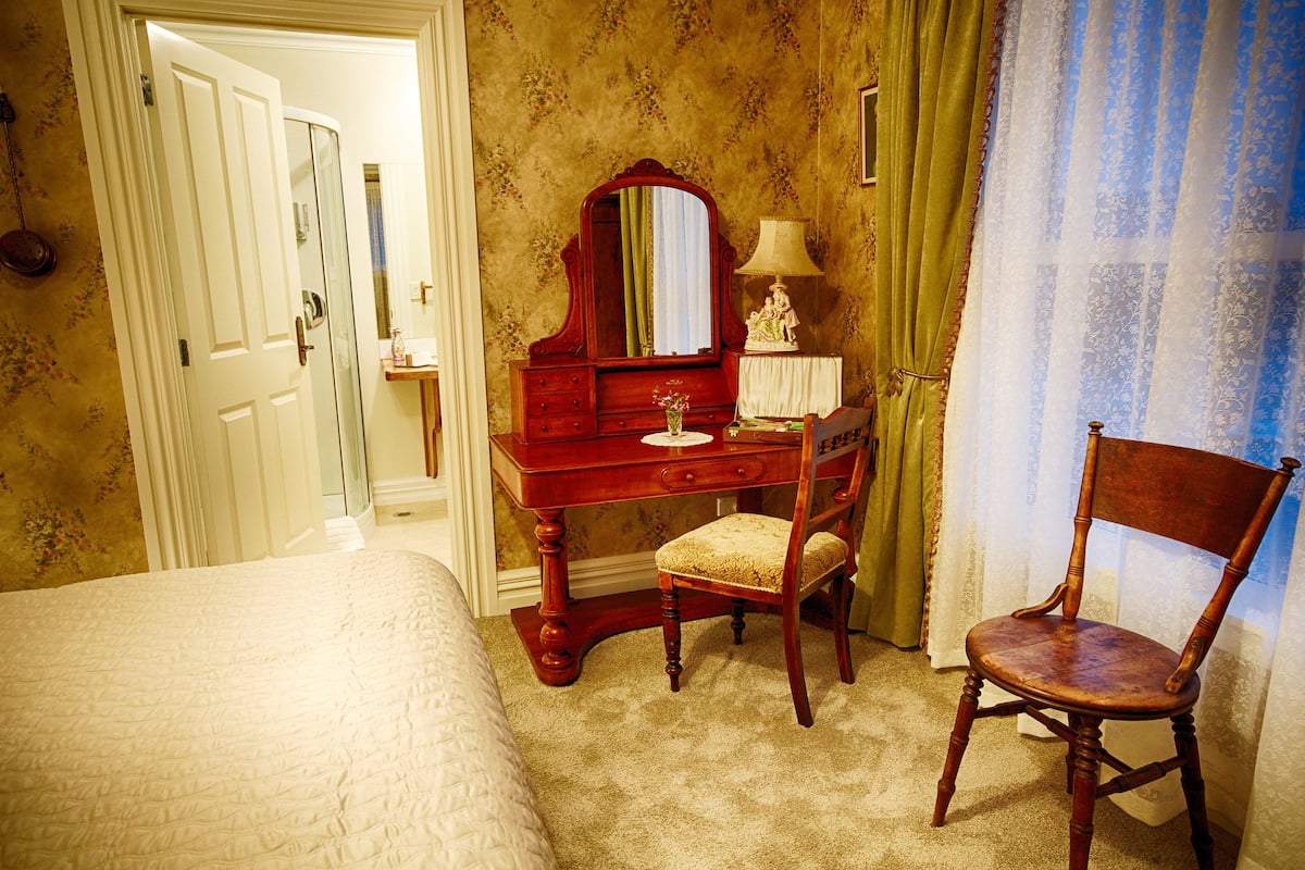 Un dormitorio con una cama, un tocador de madera con espejo, una silla y una mesita de noche con lámpara.