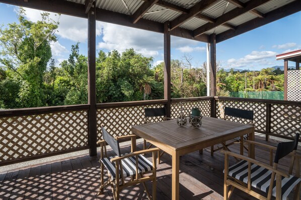 A covered outdoor seating area with a wooden table and chairs, surrounded by a lattice fence, overlooking a lush garden.