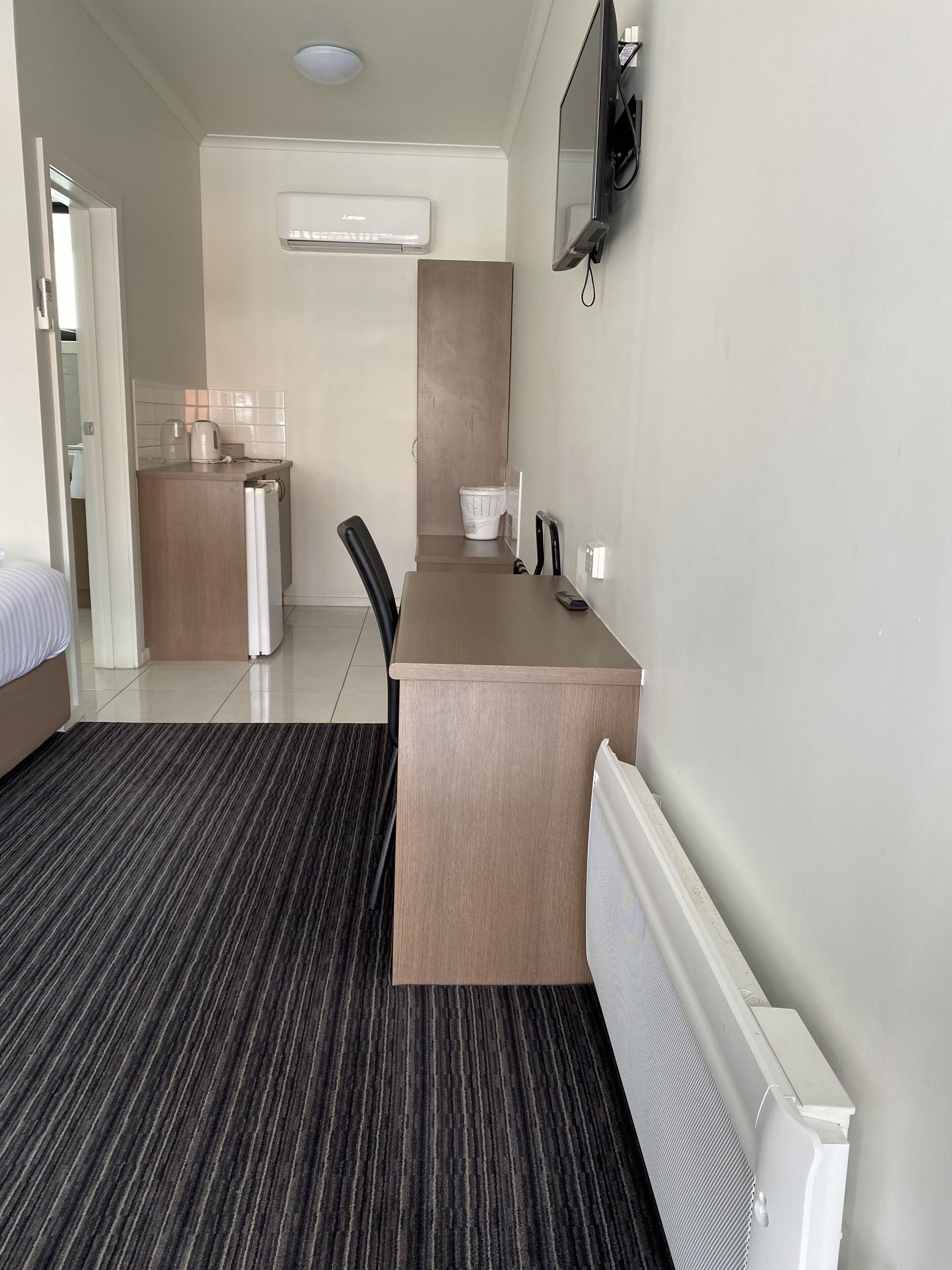 A hotel room with a bed, a desk, a chair, a radiator, an air conditioning unit, and a television.