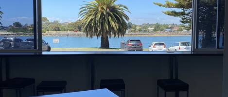 A conference room with a large table and four stools, a ceiling light fixture, and a view of a beach with parked cars and a palm tree.