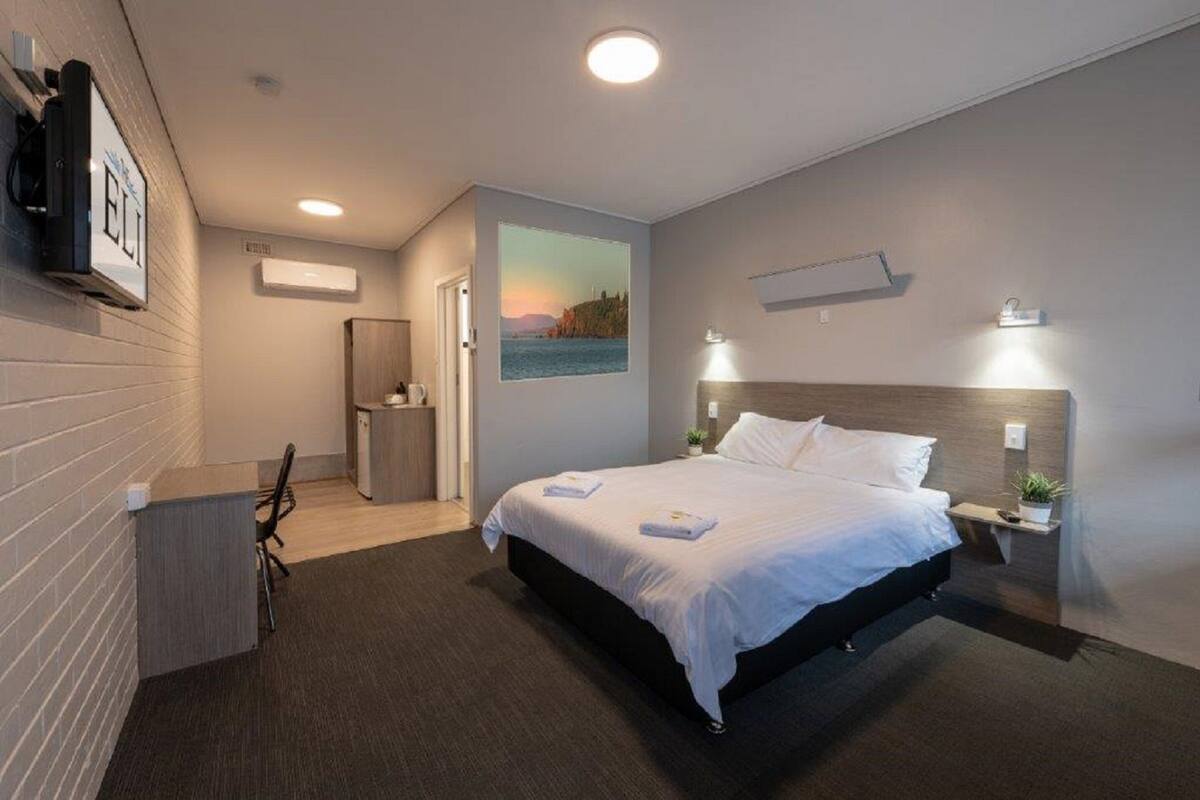 A modern hotel room with a large bed, a desk, a chair, a small kitchenette, and a painting of a sunset.