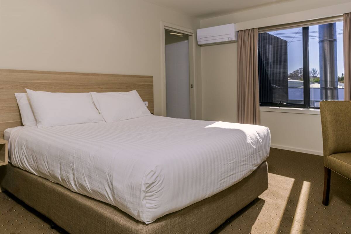 A hotel room with a large bed, a chair, an air conditioning unit, and a window with a view.