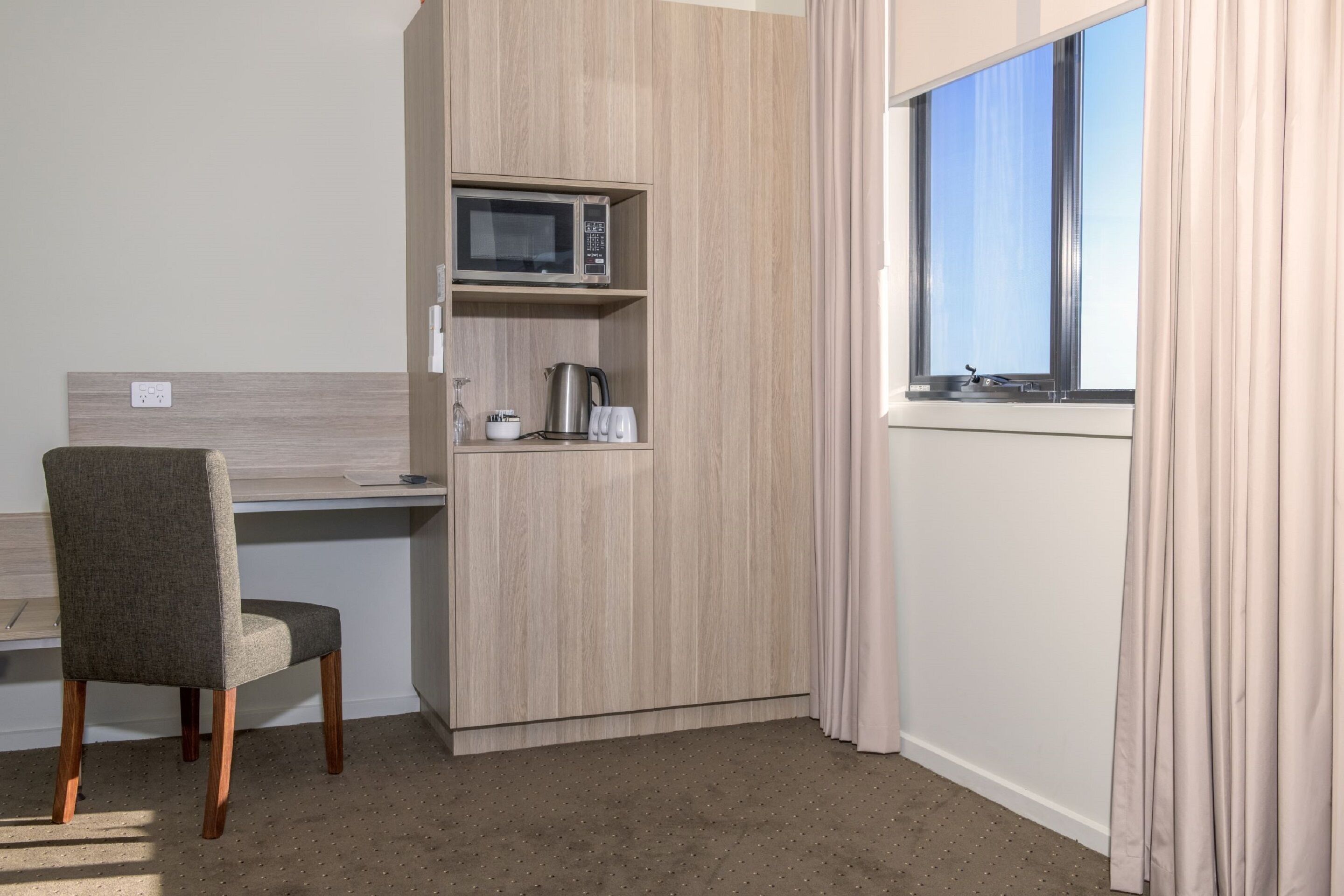 A compact hotel room with a kitchenette, a single chair, a window with curtains, and a light-colored carpet.