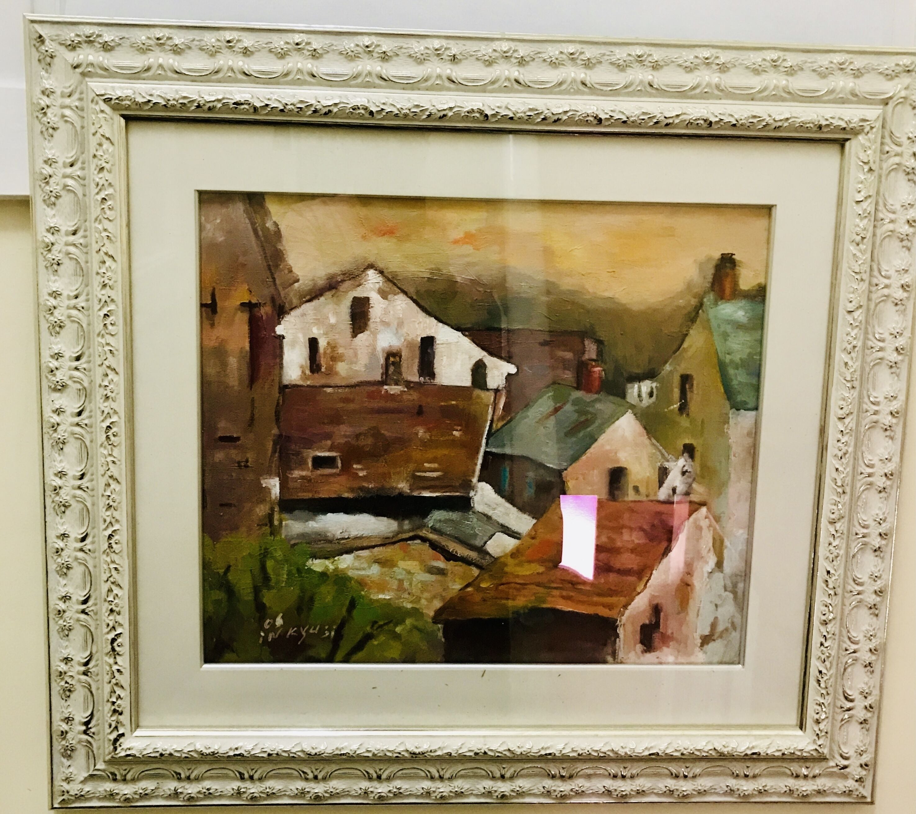 A framed painting of a rustic building with a white facade and dark roof, surrounded by greenery.