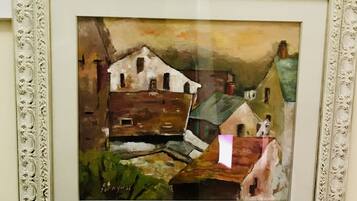 A framed painting of a rustic building with a white facade and dark roof, surrounded by greenery.