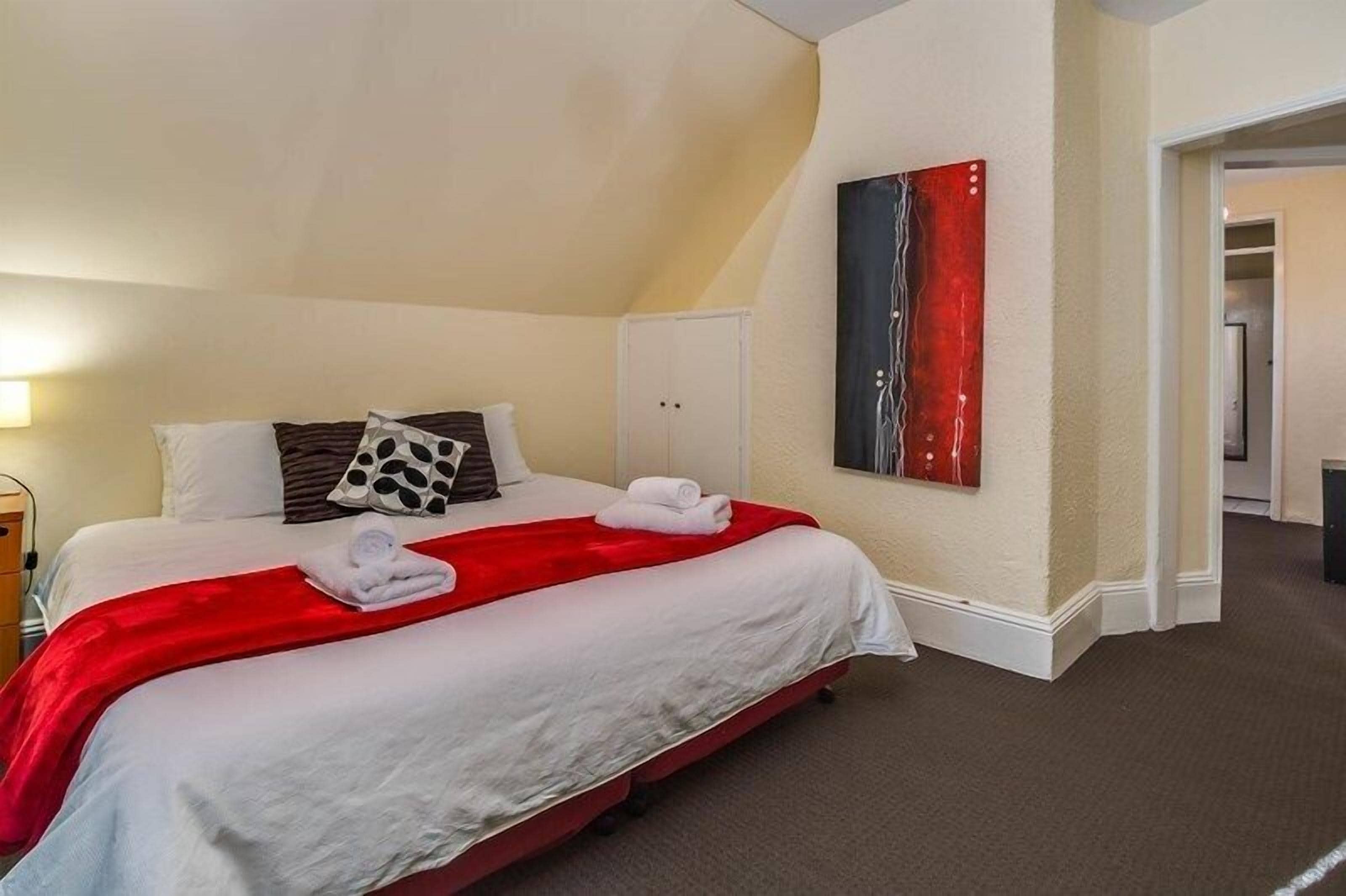 A hotel room with a bed, white bedding, a red throw blanket, and a painting on the wall.