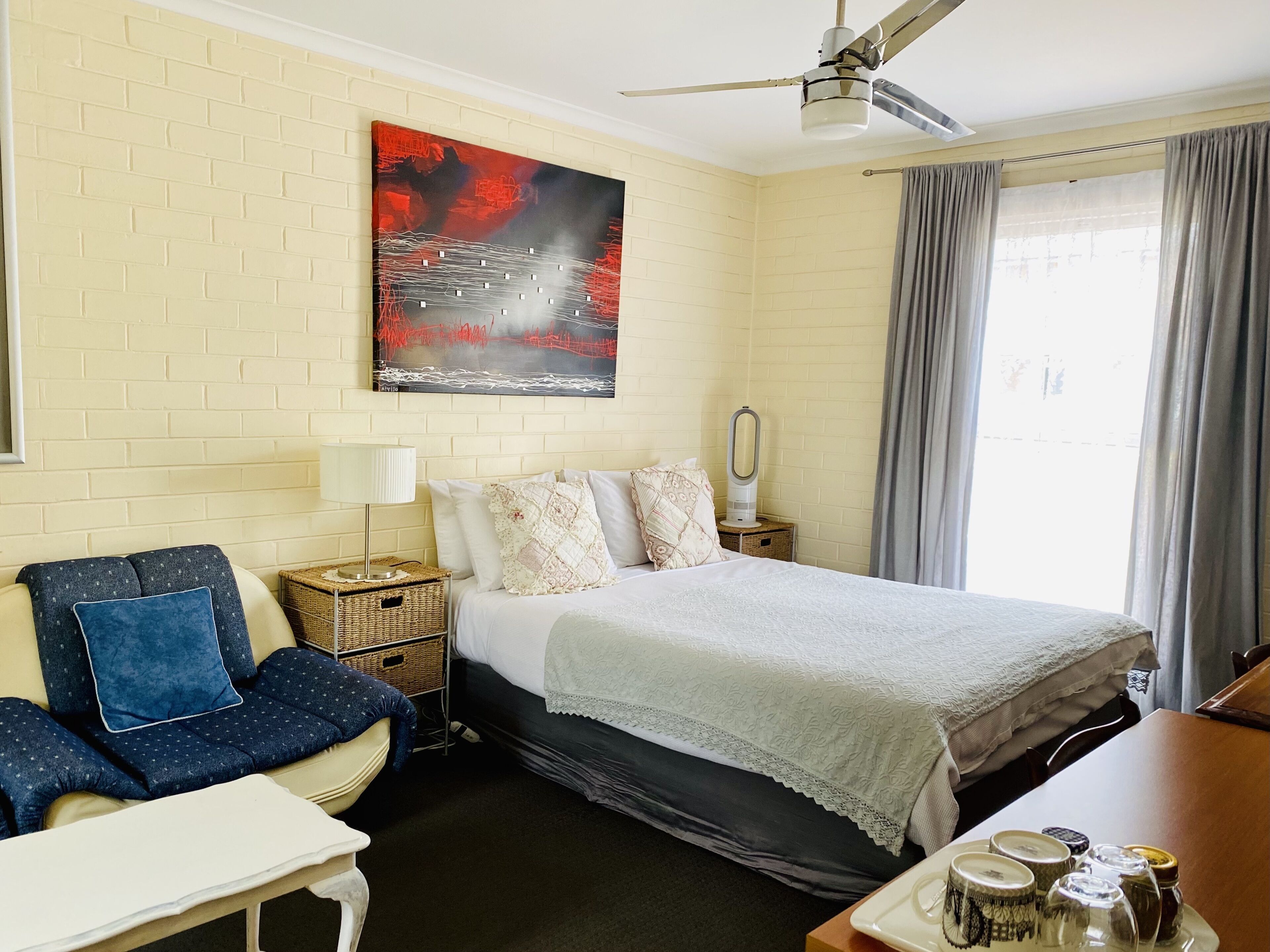 A hotel room with a bed, a chair, a table, a nightstand, a ceiling fan, a window with curtains, and a painting on the wall.