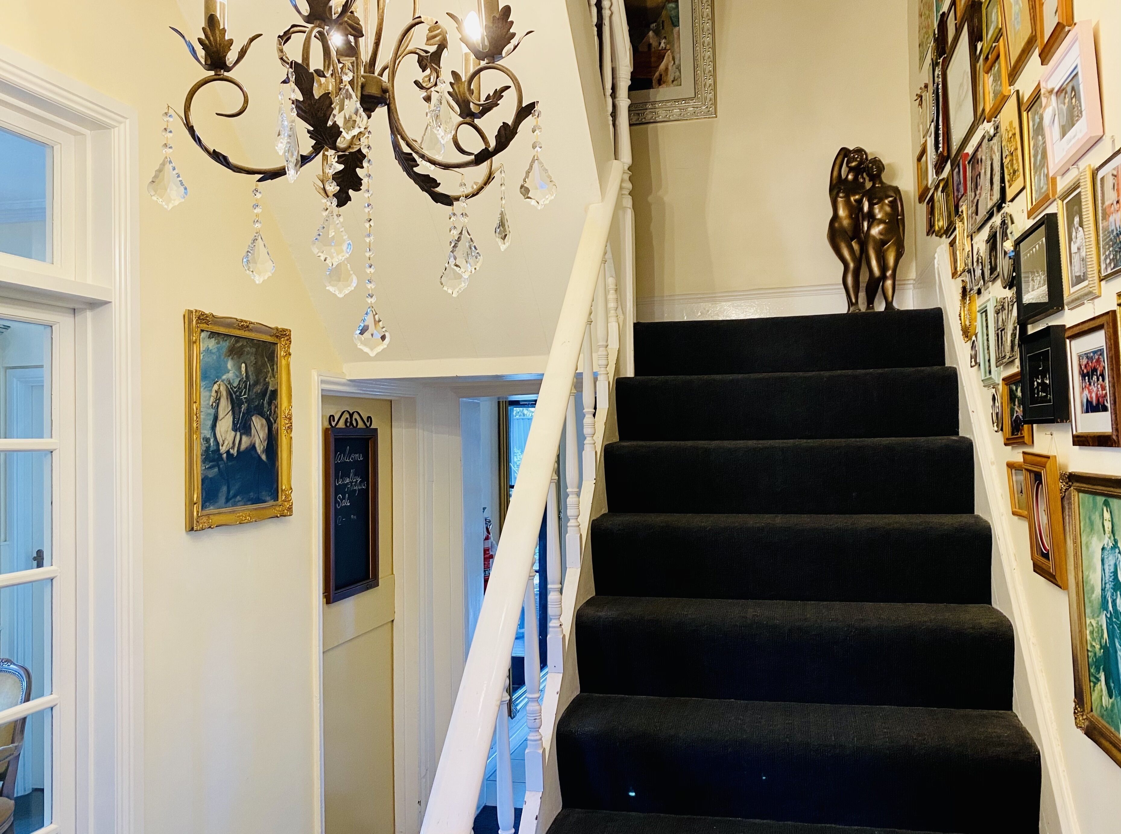 Un escalier dont les marches sont recouvertes de moquette noire, un lustre orné de décorations en cristal et un mur décoré de tableaux encadrés.