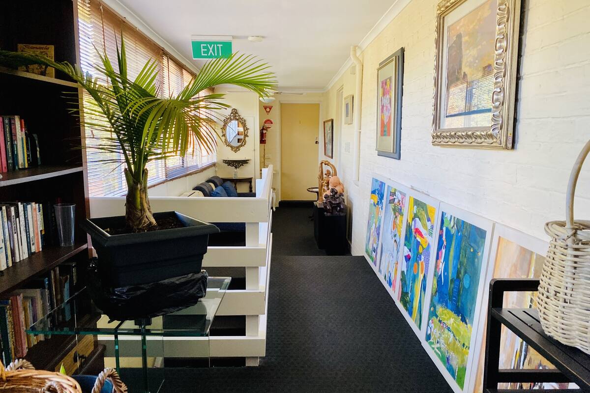 A hallway with a bookshelf, a potted plant, a white bench, and colorful paintings on the wall.