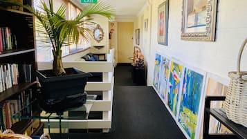 A hallway with a bookshelf, a potted plant, a white bench, and colorful paintings on the wall.