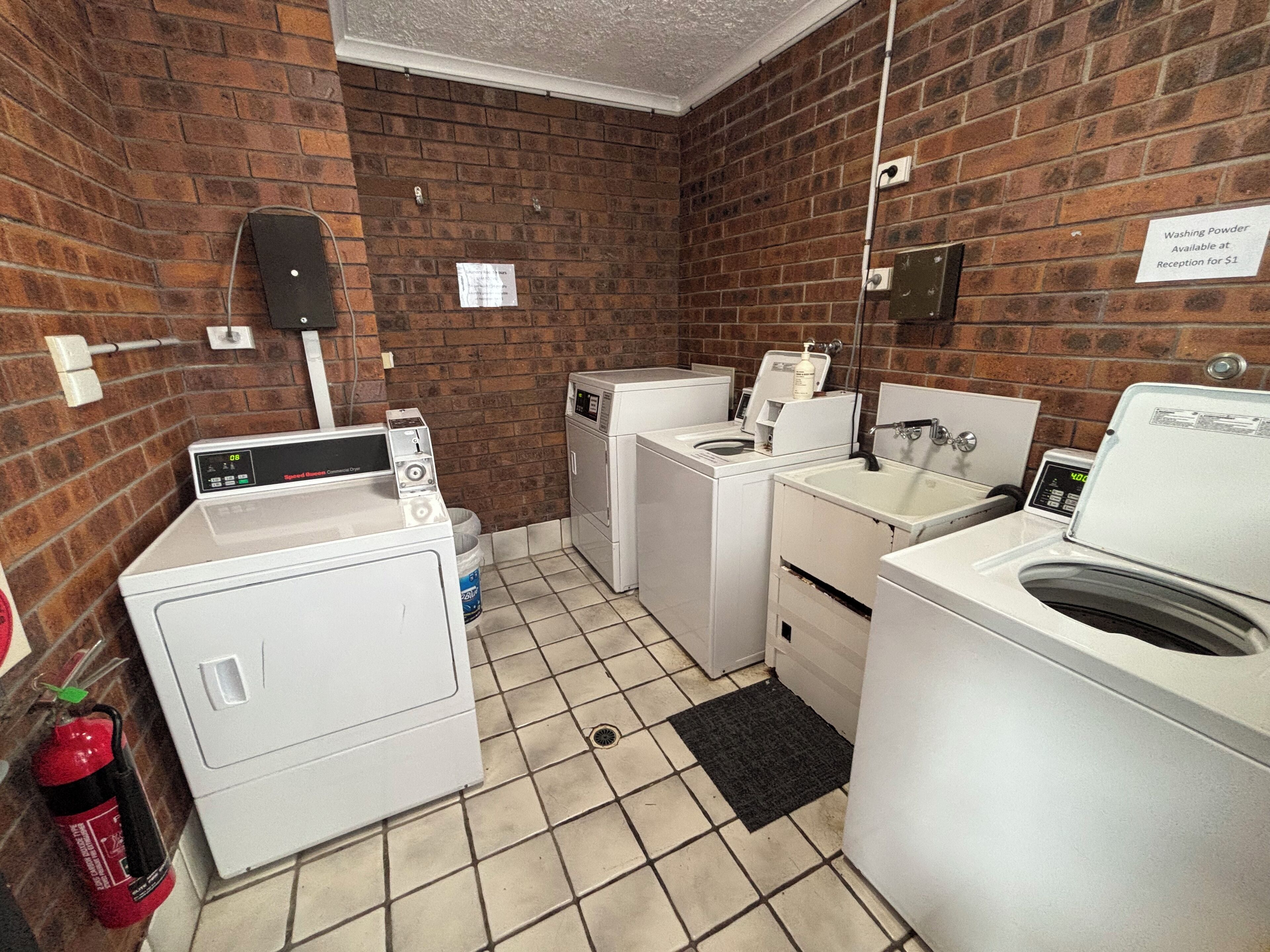 A laundry facility with multiple washing machines and dryers, a fire extinguisher, and a sign indicating the availability of washing powder for $1 at reception.