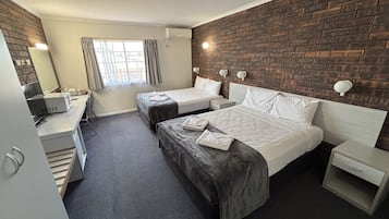 Quarto de hotel com duas camas, uma escrivaninha com computador, uma janela com cortinas e uma parede de tijolos.